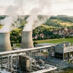Geothermie-Heizwerk mit dampfenden Kühltürmen und Rohrleitungen vor einer bayerischen Kleinstadt mit Kirchturm